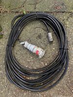 Coiled heavy duty black extension cord with industrial plug and socket ends resting on gravel ground.