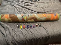 Rolled Hot Wheels play rug laid out with eight small toy cars in front including white, yellow, red, purple, blue and black models