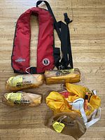 Photo of red Mustang automatic inflation life jacket with black shoulder strap and metal buckle, along with four small adult/child life jackets wrapped in yellow bags.
