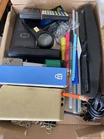 Box containing two staplers, various hole punches, scientific calculators, pencil sharpener, pens, markers, and clips.