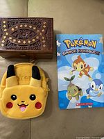 Photo showing the carved wooden box, yellow plush Pikachu coin purse, and Pokémon Sinnoh Handbook book on a table.