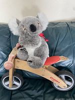 Side view of a wooden Runners balance bike with a red seat and black/gray wheels, shown with a stuffed koala on it for scale.