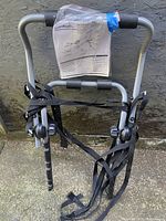 Full view of the folded SportRack bike rack showing the main frame, protective foam padding on bars, attached black securing straps, and instructions packet taped to top.