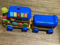 Front and side view of colorful plastic toy train showing interactive buttons and rotating gears, and attached passenger car.