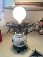 Front view of vintage table lamp with round bulb, white ceramic base with floral patterns, and metal filigree shade holder.