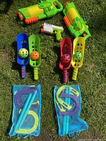 Photo showing two large green water shooters, a small green and white water gun, four colorful plastic scoops each holding a perforated ball, and horseshoe game equipment contained in two blue mesh bags.
