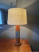 Full view of the table lamp showing the light beige textured drum lampshade and teak wood base with steel band.