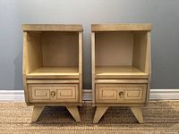 Front view of two vintage nightstands side by side showing open upper shelves, single drawers with carved geometric fronts and brass knobs, and tapered legs.