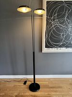 Full view of lamp turned on against a gray wall showing two illuminated heads and foot pedal control on hardwood floor