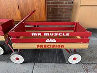 Side angle image of the red wooden Mr Muscle toy wagon showing white text stamped on wood and the wheel structure.