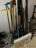 Garden tools including post hole digger, rakes, brooms, shovel, and miscellaneous tools leaning against the wall.