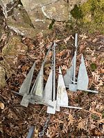 Two galvanized steel boat anchors with attached chains resting on dry leaves and stone ground outdoors.