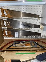 Hand saws and saw blades mounted on pegboard, wooden handles with metal blades shown, including Disston brand.