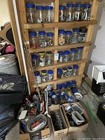 Wide view of a wooden rack holding multiple clear plastic jars with blue lids filled with assorted screws, bolts, nuts, washers, and hardware parts, along with additional boxes of hardware on the floor.
