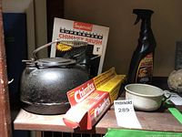 Shows cast iron kettle, Imperial chimney brush, chimney cleaning rods boxes, spray bottle and metal cup together on table.