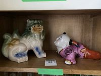 Shelf displaying two ceramic figurines: a foo dog and a reclining figure, showcasing their size and relative scale.