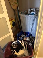 Wide image showing broom, step ladder, ironing board, plastic bin with assorted cleaning items and tools inside, mechanical scale, and plumbing or handheld tools in the bin.