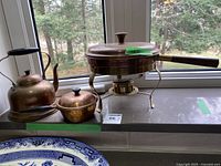 Copper chafing dish, small copper pot with cover, and teapot on a countertop by window