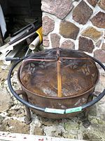 Full view of the round metal fire pit with mesh cover placed outdoors on stone flooring, showing rust and decorative cutouts.