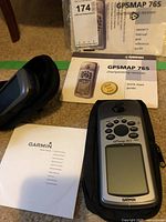 Photo showing the Garmin GPSMAP 76S device, owner's manual, quick start guide, and protective case on a carpeted floor.