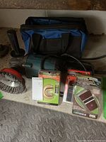 Power tool with side handle attached, wire brush on front, power cord, blue and black carry bag in background, packaged blades in front