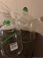 Two clear glass bell-shaped domes of different sizes, 10 inches and 13 inches high, placed on a countertop with green tape labels on knobs