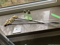 Set of six metal meat skewers with brass animal and human finials alongside the pair of green Depression glass salt and pepper shakers on a stone windowsill.
