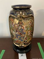 Full frontal photo of the tall ceramic vase on black stand showing the detailed hand-painted figures and gold accents on a dark background.