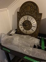 Full view of the vintage brass clock face showing the Roman numerals, clock hands and decorative embossed brass housing. Wrapped pendulum and weights are visible below the clock on a black surface.