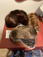 Photo of three hats including a dark brown round fur hat, a light brown fur cylindrical item, and a brown leather aviator-style hat with fur trim and ear flaps on a red surface.