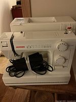 Full front view of the white Janome HD-1000 sewing machine with foot pedal wrapped with power cord.