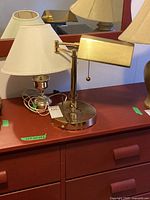 Desk lamp with brass adjustable arm and rectangular shade, with pull-chain switch, placed on a reddish-brown wooden surface in front of a mirror.