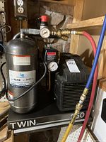 Front view of LE Twin air compressor showing the main silver tank with LE decal, pressure gauges, attached red and blue hoses with brass fittings, and black motor housing.