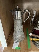 Photo showing lemonade jug with silver plated top and detailed glass base, lid closed, on wooden shelf