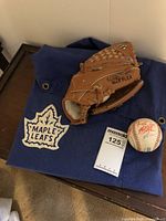 Max Flex baseball glove placed on top of folded blue Maple Leafs cloth with white logo, accompanied by a signed baseball and a MaxSold lot number card.