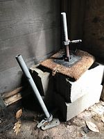 Two metal support jacks with threaded adjustable rods and flat bases with bolt holes, placed near and on concrete blocks with carpet, showing visible surface rust.