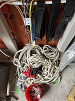 View of coiled white ropes, metal chains, and red pulleys on the floor, with climbing or safety straps hanging above.