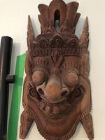 Close-up front view of a hand-carved wooden mythical creature face with detailed ornamental headgear, mounted on a wall.