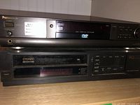 Panasonic DVD-A310 DVD/CD player stacked on top of RCA stereo VHS VCR on a wooden surface, showing front panel features like power, display, and slot for VHS tape.