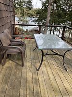 Patio glass top table with four matching chairs arranged on outdoor wooden deck, lake and trees seen in background.