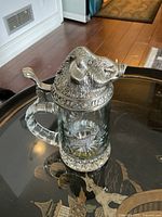 Overall view of the glass beer stein with pewter boar head lid sitting on a black table.