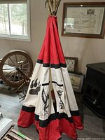 Front view of teepee showing the red and white fabric with black tribal animal designs, standing indoors next to a spinning wheel and framed pictures.