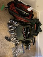 Red and green fabric tool bags open on floor showing various tools including sockets, bit sets, wrenches and handles.