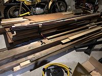 Long mixed lumber pieces stacked horizontally with various widths and lengths visible, some with clamps attached, placed on a concrete floor with motorcycle in background.
