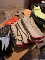 Multiple pairs of beige leather work gloves with red trim cuffs and other gloves stacked on table.
