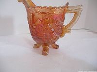 Side view of the orange carnival glass creamer showing the handle, spout, scalloped rim and raised Thorn and Thistle decorative pattern with iridescent finish.