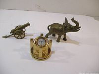 Three brass items on white surface: elephant figurine, small round clock in geometric stand, and decorative cannon.