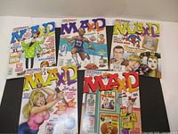 Five MAD magazines displayed spread out on a surface, showing various colorful comic-style covers with caricatures and parodies.