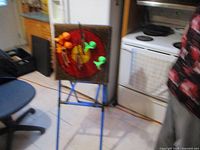 Standing target board for axe and dart throwing game with orange and green plastic axes and darts stuck in the board, mounted on a blue metal tripod stand