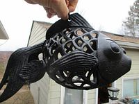 Black cast iron fish outdoor decoration being held by hand, showing hollow body with cutout scales and textured fins and tail.